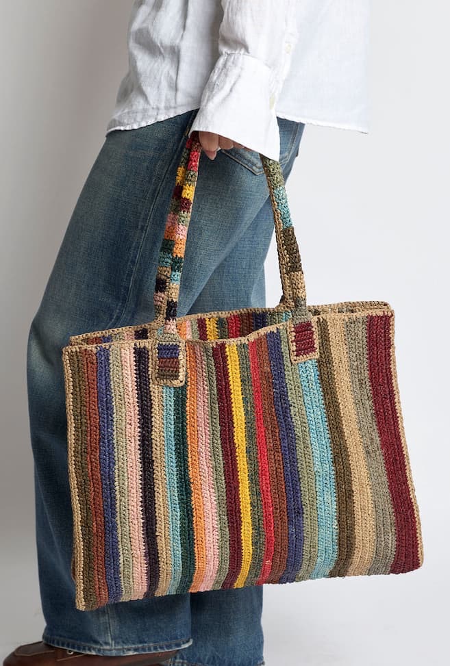 A colorful handwoven tote bag with vertical stripes in shades of blue, red, yellow, green, pink, and natural tan is carried by a person wearing a white shirt and blue jeans. The textured raffia-style bag features sturdy woven handles and a spacious rectangular shape, blending artisanal craftsmanship with everyday practicality for shopping, travel, or casual outings.