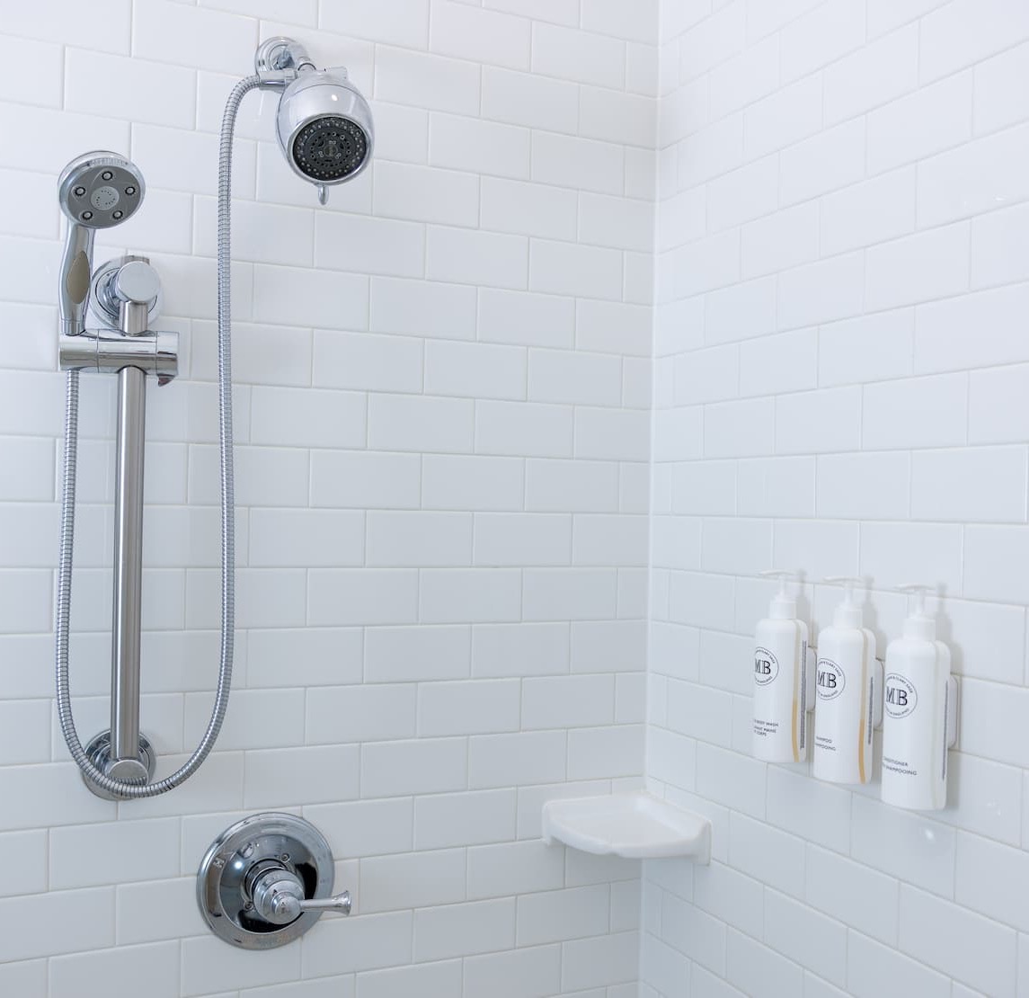 Minimalist shower with white subway tile walls, chrome shower fixtures, and three mounted pump bottles for body wash, shampoo, and conditioner on a corner shelf.