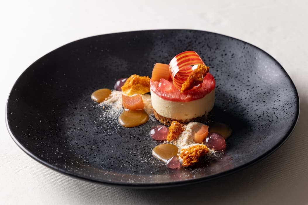 An elegant plated dessert is presented in a matte black bowl, featuring a small round cheesecake or mousse atop a crisp base. The dessert is garnished with delicate ribbons of pink rhubarb, caramel-colored sauce, translucent fruit gel pearls, crunchy honeycomb-like shards, and a dusting of fine crumbs. The dramatic contrast between the colorful components and the dark plate highlights the refined presentation and fine-dining craftsmanship.