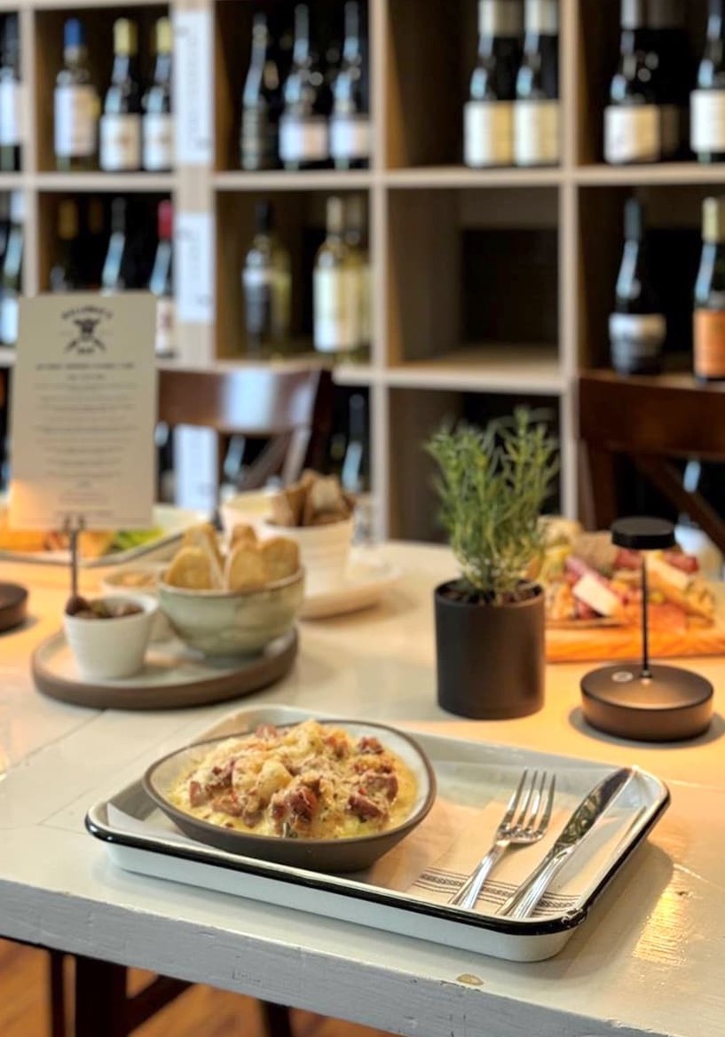 A table set with plated food, cutlery, small potted herbs, and wine glasses sits in front of shelves filled with various bottles of wine in a cozy restaurant setting. Fearrington Village