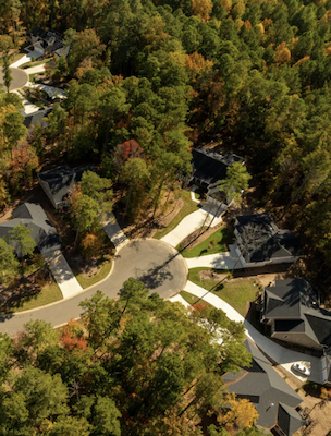 Aerial view of a wooded residential neighborhood with curved streets and several homes nestled among dense trees showing early fall colors.