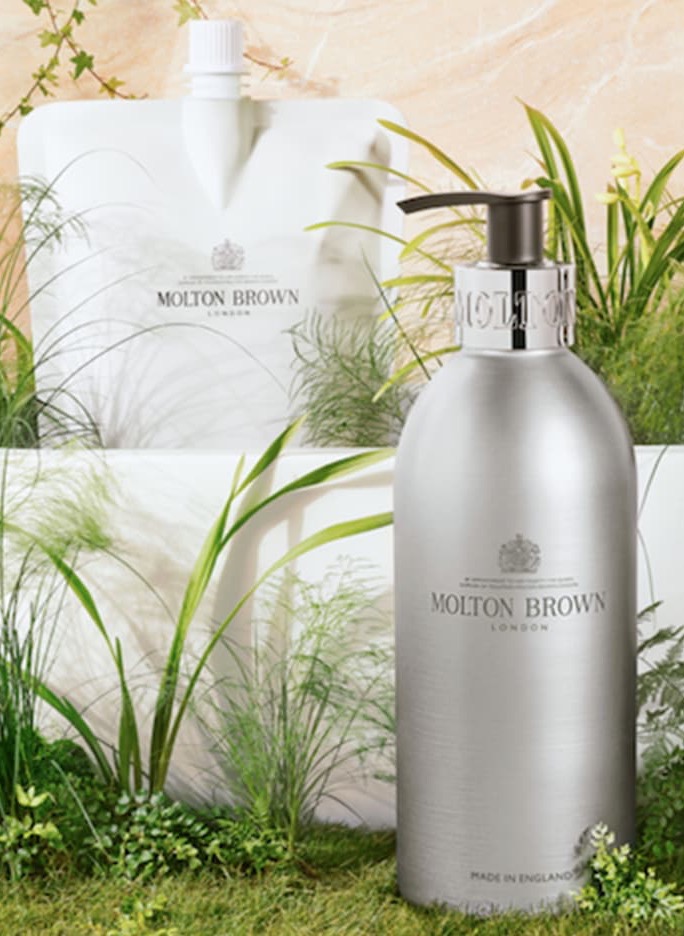 A brushed silver Molton Brown London soap dispenser stands among lush green plants, with a refill pouch visible behind it. The product is styled in a natural, spa-like setting, emphasizing clean lines and a fresh, botanical feel.