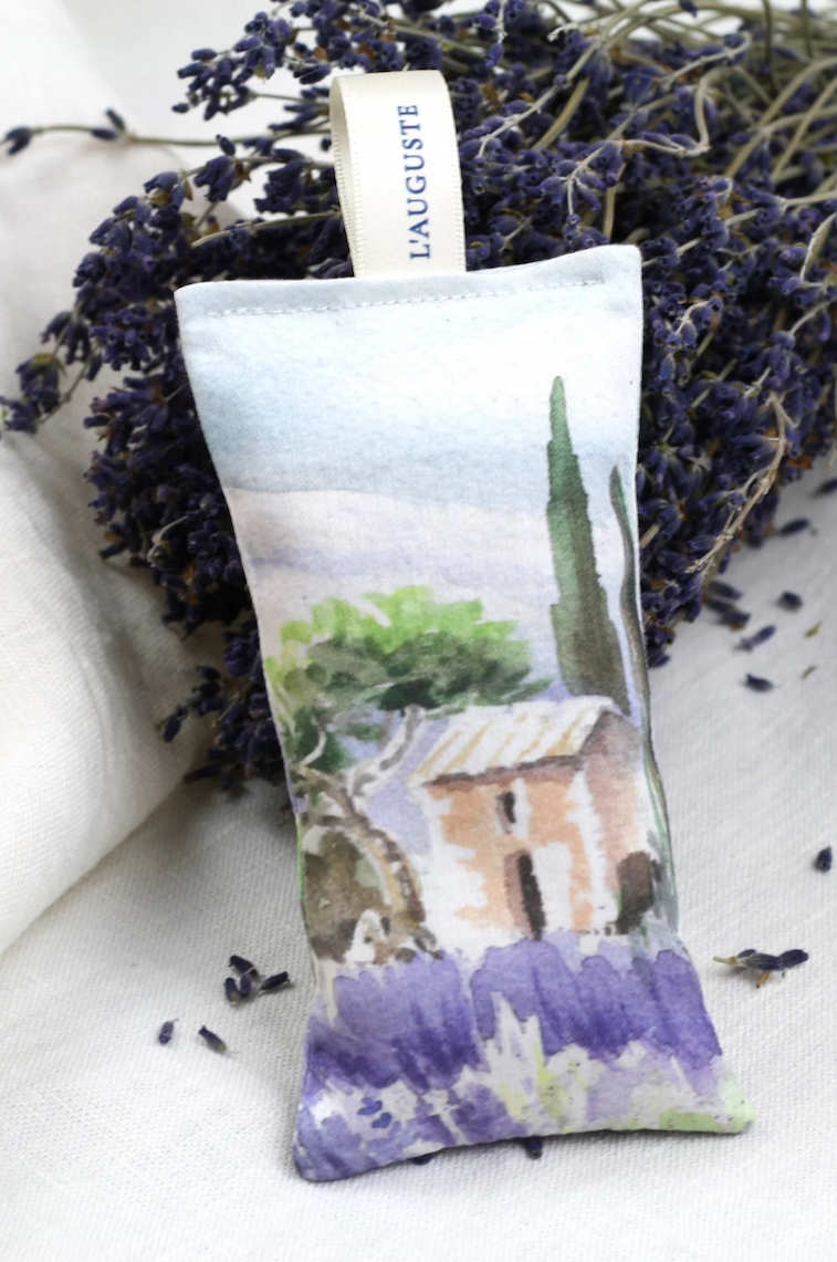 A lavender sachet with a soft watercolor illustration rests against a bundle of dried lavender, with loose petals scattered on a light fabric surface.