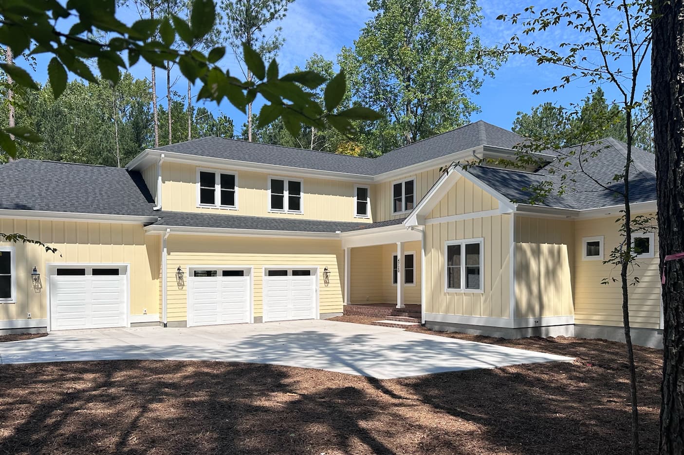 Newly built pale yellow home with white trim, three-car garage, and a welcoming covered entry surrounded by mature trees. Fearrington Village.