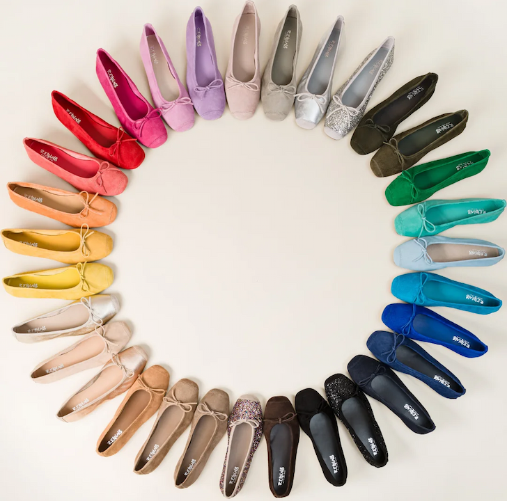 A full spectrum of suede ballet flats arranged in a circle on a light background, showing dozens of colors from red and pink to blue, green, gray, and black.