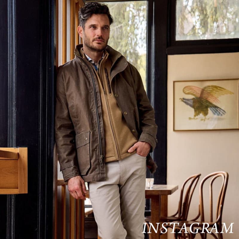 A man stands indoors by a window, wearing a brown jacket, tan sweater, and beige pants. He rests one hand in his pocket. Behind him are wooden chairs, a table, and a framed bird illustration on the wall. Fearrington Village