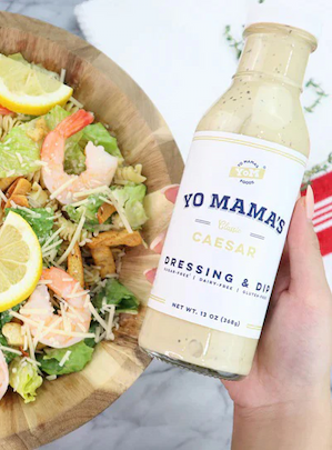 Hand holding a bottle of Yo Mama’s Caesar dressing beside a wooden bowl of Caesar salad with shrimp, lemon wedges, and grated cheese.