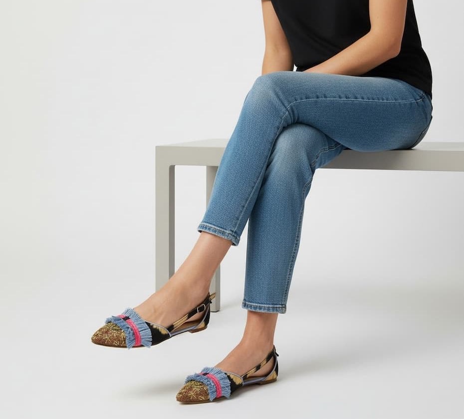 Studio image of a person seated on a minimalist white bench wearing a black top and cropped light-wash jeans. They are styled with pointed-toe slingback flats featuring a metallic gold woven pattern, black trim, and playful blue fringe bands accented with bright pink stripes across the toes. The colorful details add a whimsical, statement-making touch to an otherwise casual look.