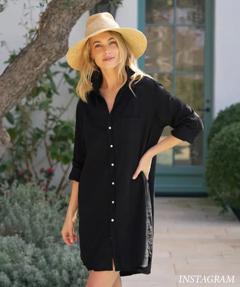 A woman in a relaxed black shirt dress stands outdoors in a sunlit garden setting, wearing a wide-brim straw hat and posing casually near a tree and a green door.