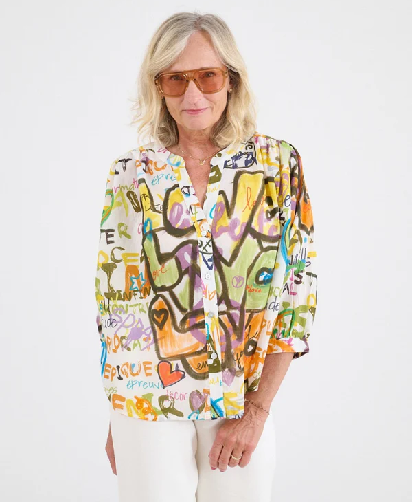 Woman wearing amber-tinted sunglasses and a colorful, graphic-print blouse with abstract lettering and heart motifs, paired with light pants against a neutral background.