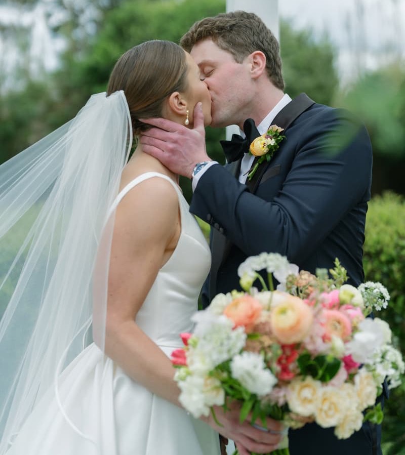Bride and groom share a kiss in the gardens, with the bride holding a colorful bouquet of spring flowers. Fearrington Village.