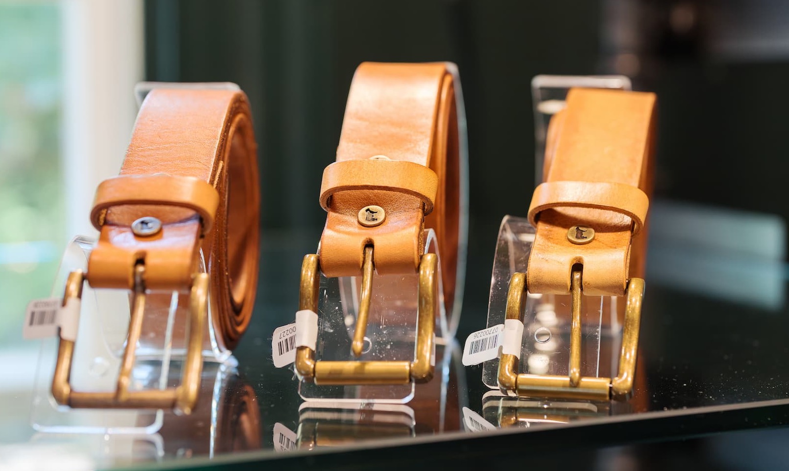 Three tan leather belts displayed on clear stands, each with a brass buckle and clean, minimal design. The polished hardware and smooth leather create a classic, understated look, with soft reflections on the glass surface.