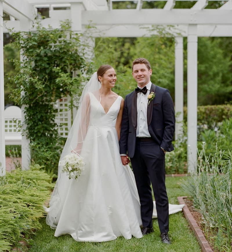 Bride and groom standing hand in hand beneath the white pergola in Fearrington Village's gardens after their ceremony, surrounded by lush greenery and spring blooms.