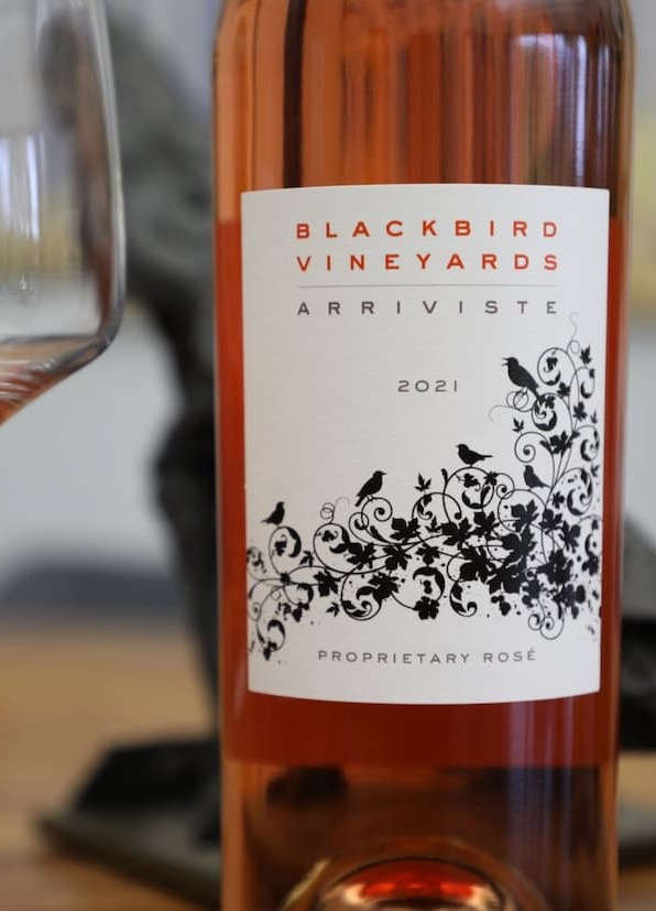 Close-up of a bottle of Blackbird Vineyards Arriviste 2021 Proprietary Rosé beside a partially filled wine glass. The elegant white label features intricate black vine and bird illustrations, while the rosé wine glows a warm salmon-pink through the bottle. The softly blurred background keeps the focus on the wine’s distinctive packaging and color.