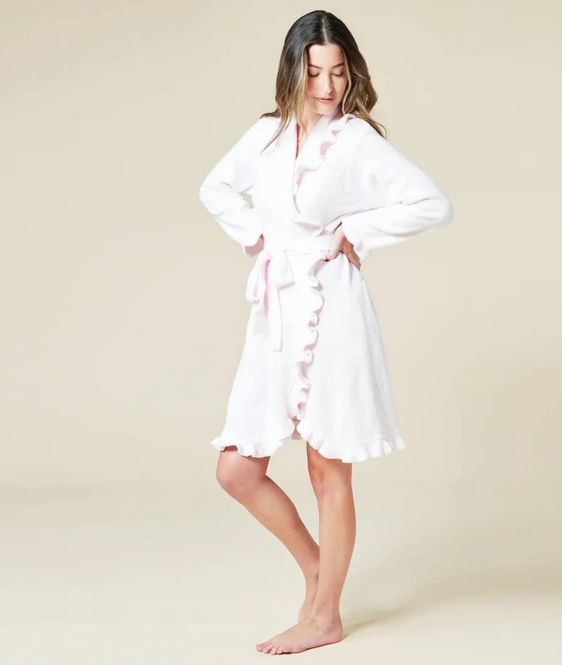 A model stands barefoot against a soft beige background wearing a plush white robe trimmed with pale pink ruffles along the collar, front opening, and hem, cinched at the waist with a matching tie belt.