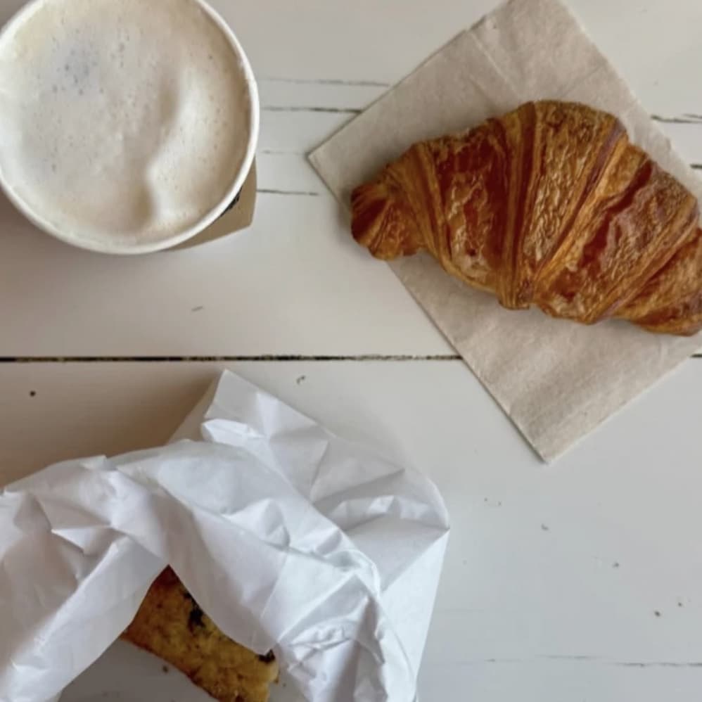 Overhead view of a cup of frothy coffee and a golden croissant on a napkin, with a partially wrapped pastry on white wooden table boards.