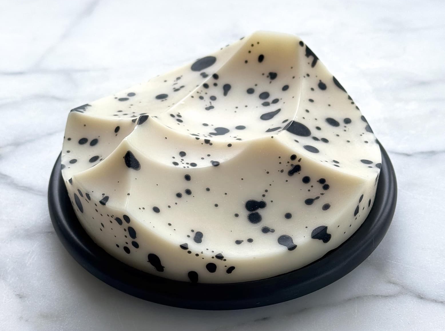 A sculpted white Ume Erode soap with black speckles resting on a round black dish against a marble surface.