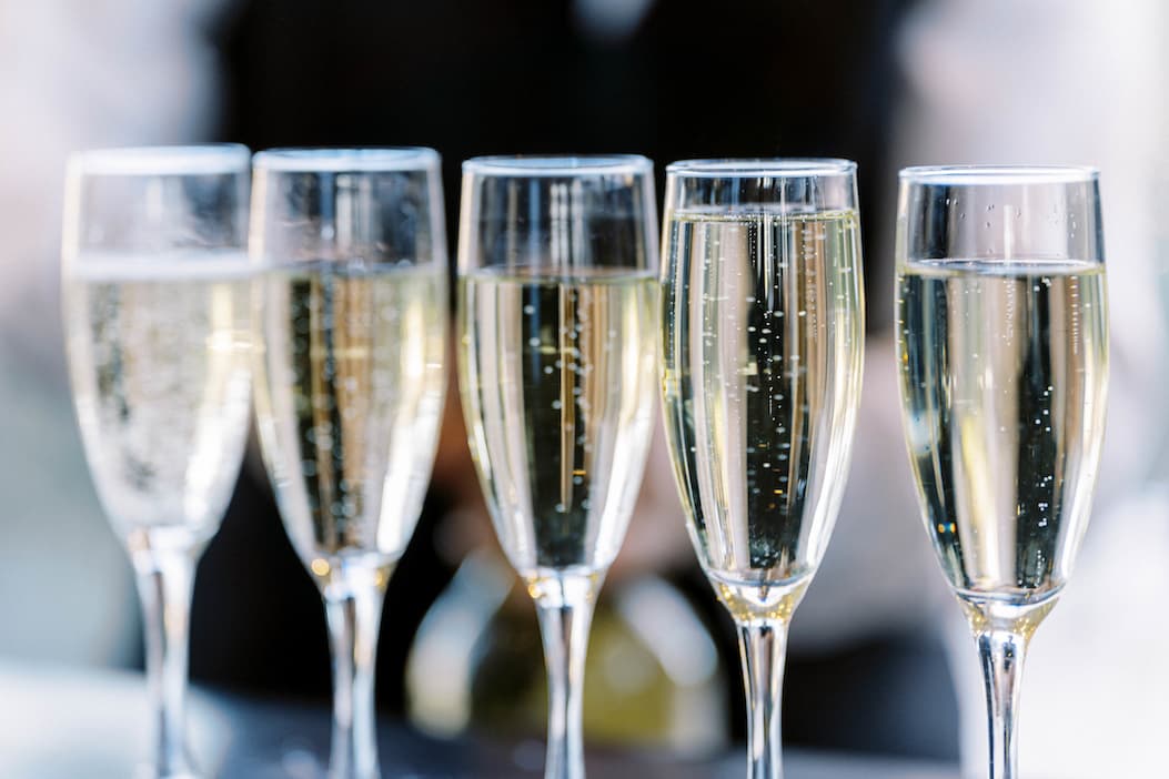 Five tall champagne flutes filled with sparkling wine are arranged in a row, with bubbles rising inside each glass. The background is softly blurred, drawing focus to the glasses. Fearrington Village