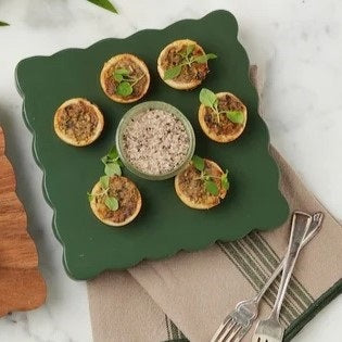 Deep green scalloped serving board topped with bite-sized appetizers arranged around a small bowl of dip. The board is styled on a neutral linen napkin with silver serving utensils, creating an elegant presentation for appetizers, cocktails, or casual entertaining.