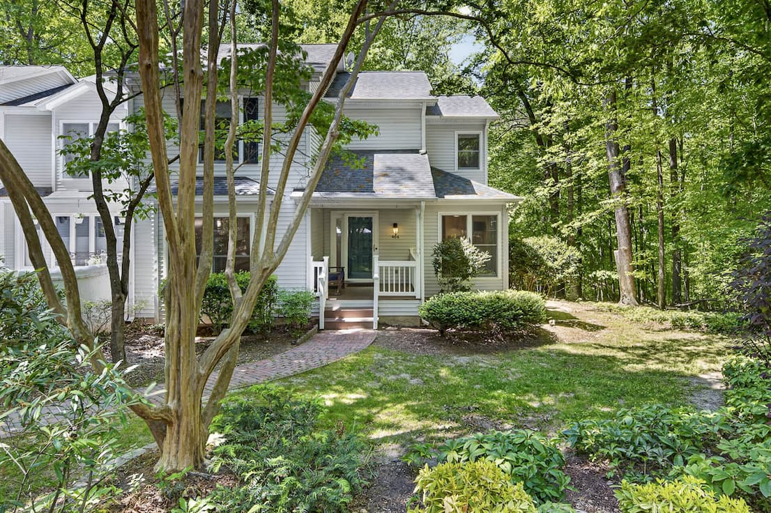A light gray, two-story home sits among mature trees, with a small covered front porch, brick walkway, and landscaped garden beds leading through a shaded, wooded yard.