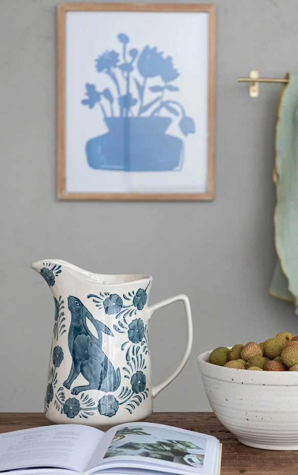 Handpainted ceramic pitcher with a blue rabbit and floral motif, sitting on a wooden table beside a bowl of fruit and an open book.
