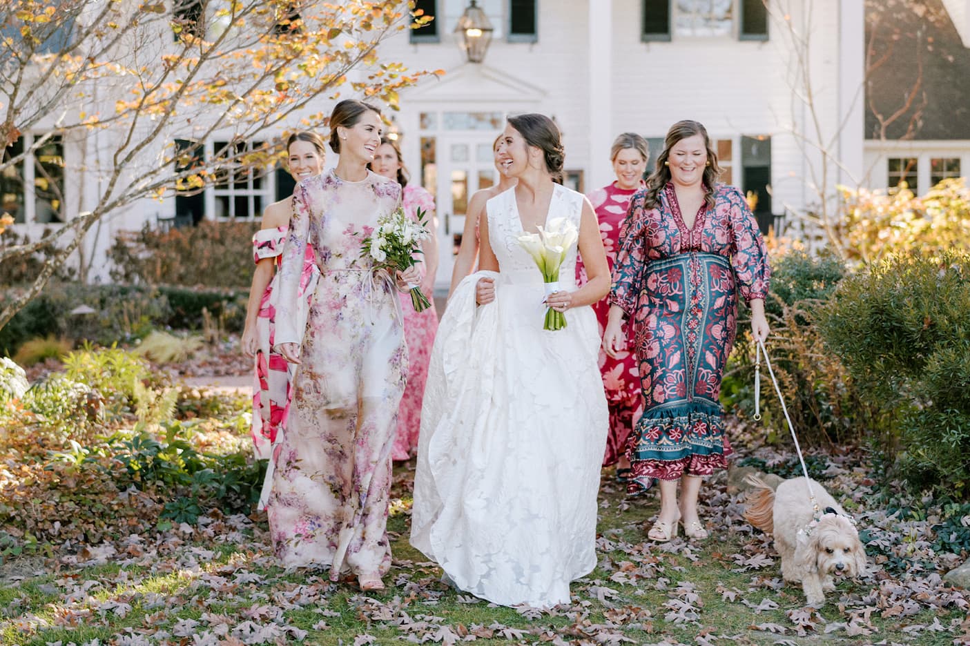 Bride in a white gown walks through a garden with bridesmaids in colorful dresses outside a white inn, while a small dog on a leash trots beside them.