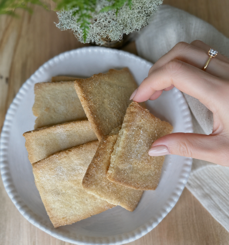A manicured hand wearing a delicate solitaire ring reaches for a sugar-topped shortbread cookie from a white ceramic bowl. Soft linens, warm wood tones, and natural textures create an elegant, cozy kitchen moment.