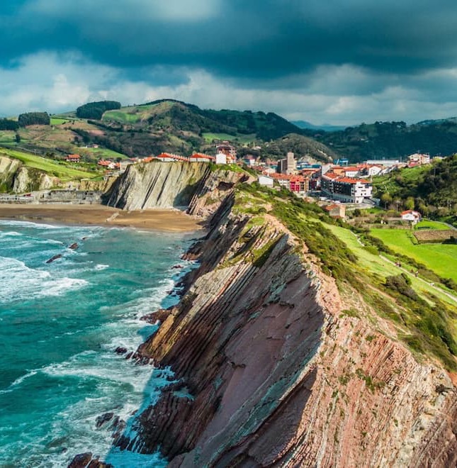 A dramatic stretch of Spain’s northern coastline features rugged, layered cliffs plunging into turquoise waters, with rolling green hills, winding walking paths, and a seaside village nestled between the mountains and the sea beneath moody, cloud-filled skies.