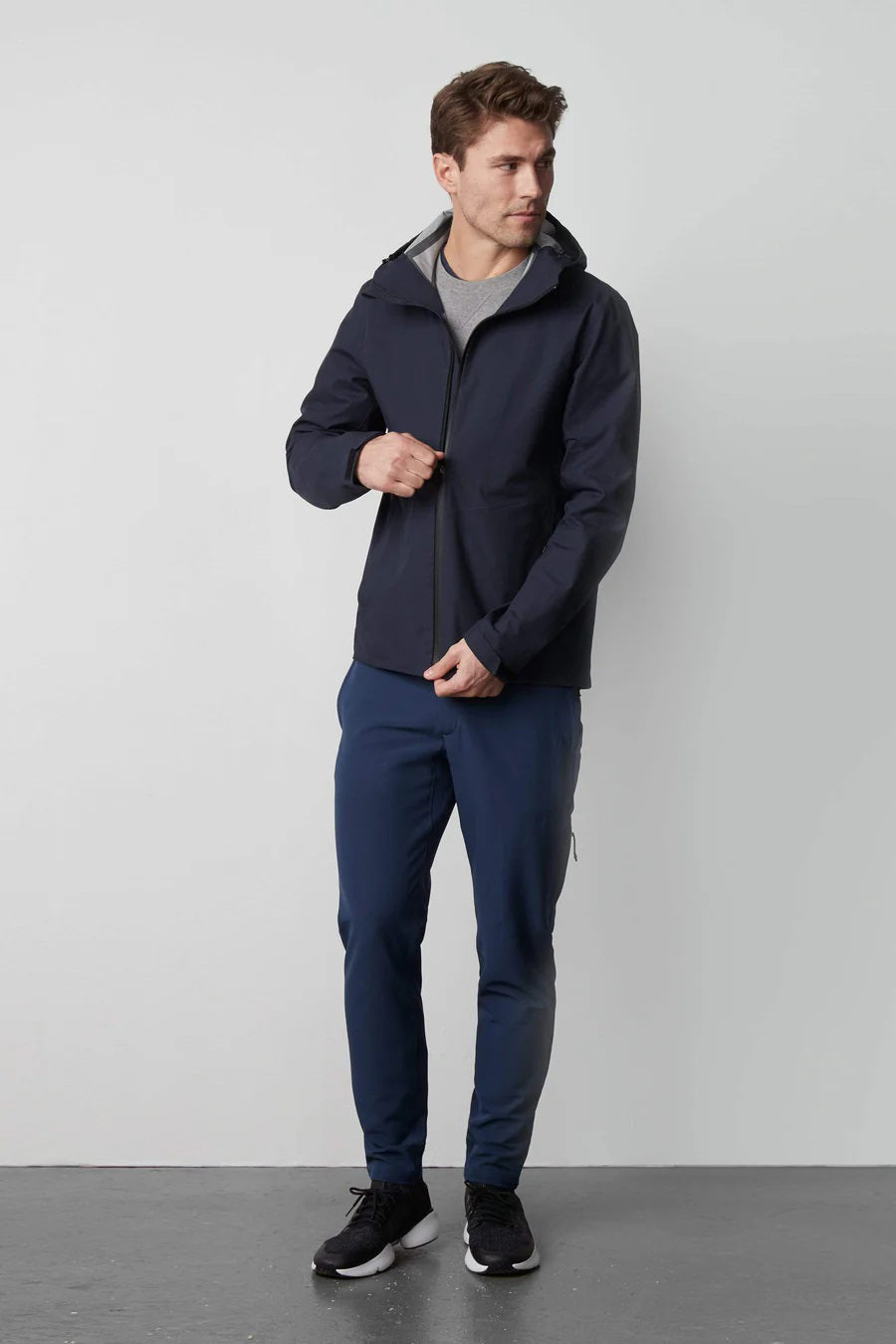 Man wearing a navy zip-front jacket with hood, blue pants, and black sneakers stands against a light gray background.