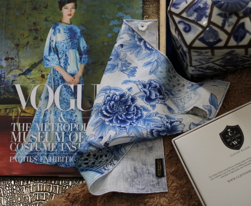 Flat lay of a blue-and-white floral pocket square alongside a Vogue magazine, a patterned ceramic jar, and a branded card, arranged on a textured surface.