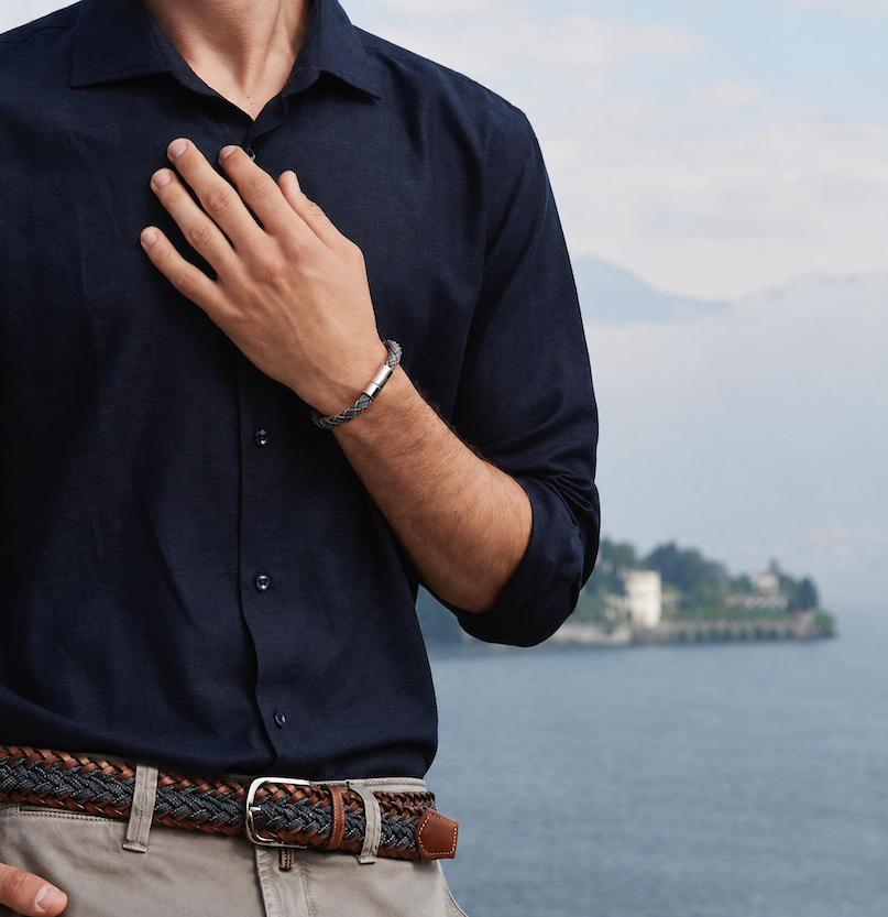 Close-up of a man in a navy button-down shirt wearing a braided bracelet with a metal clasp, hand resting at his chest, with a soft coastal landscape in the background.