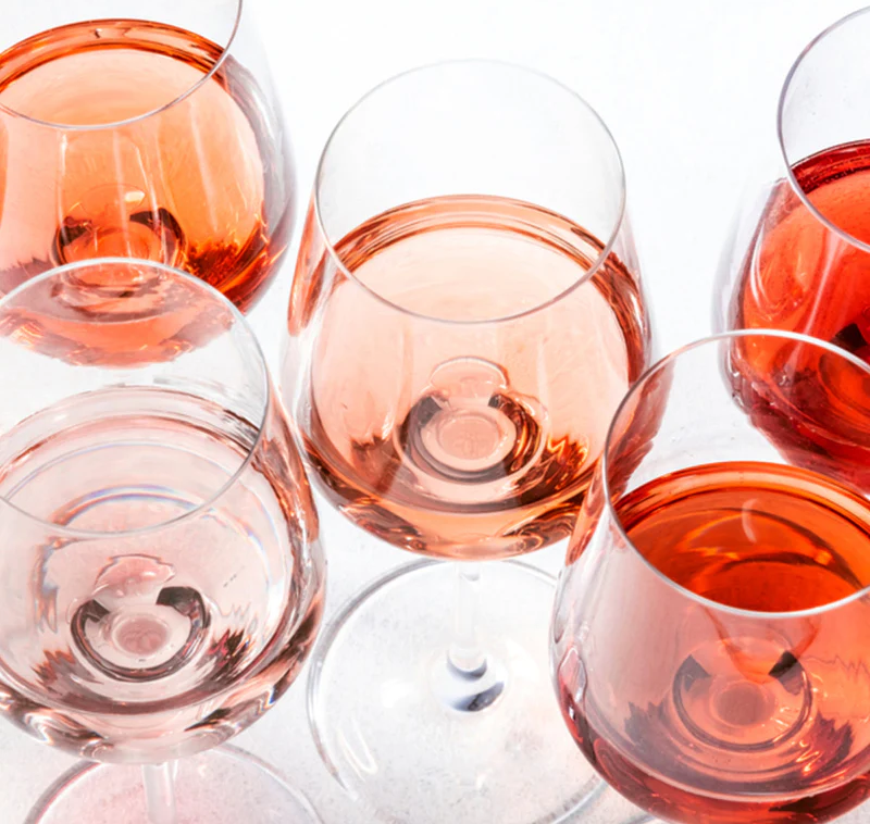 Overhead view of five wine glasses filled with rosé in varying shades from pale blush to deep pink on a light surface.
