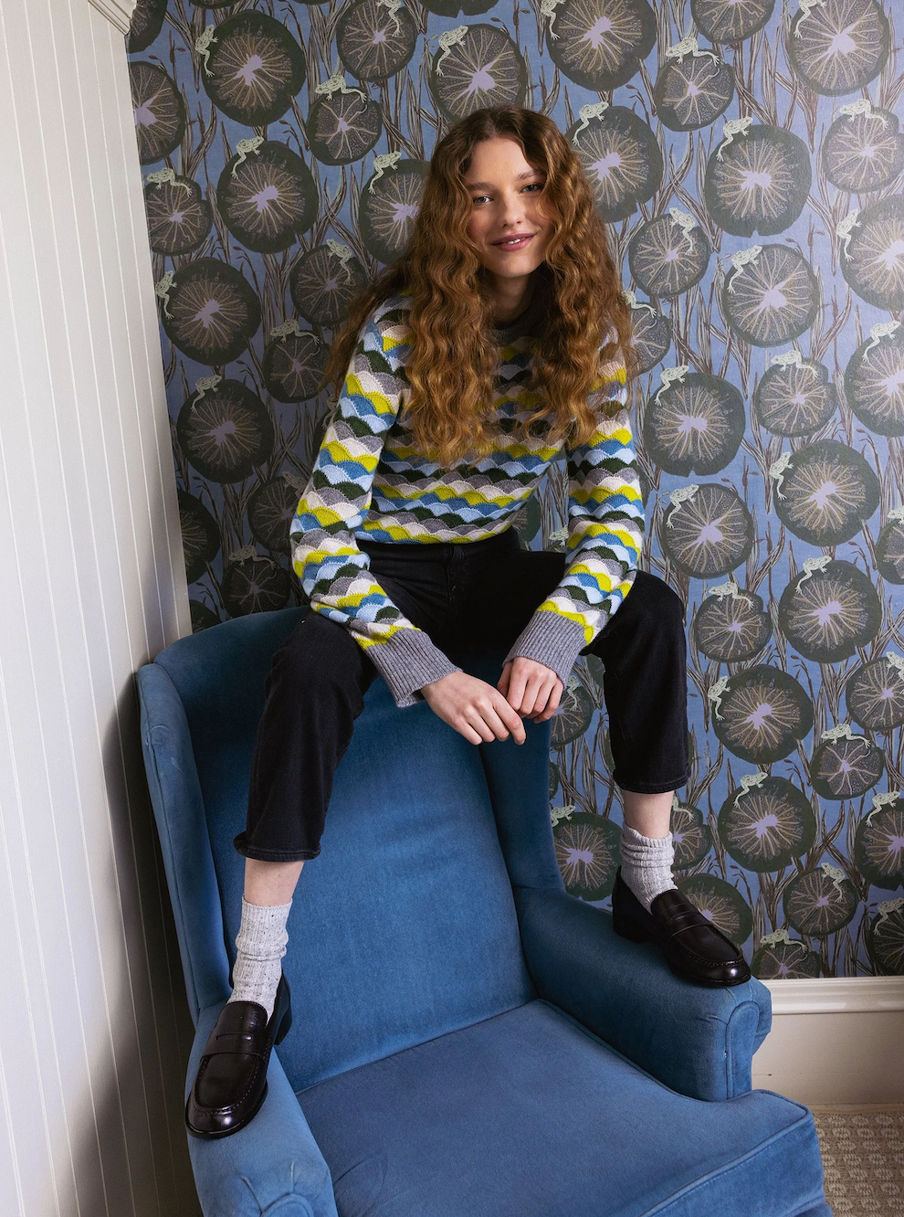 Woman wearing a colorful patterned sweater, black jeans and loafers, seated playfully on a blue armchair against botanical wallpaper. Fearrington Village.