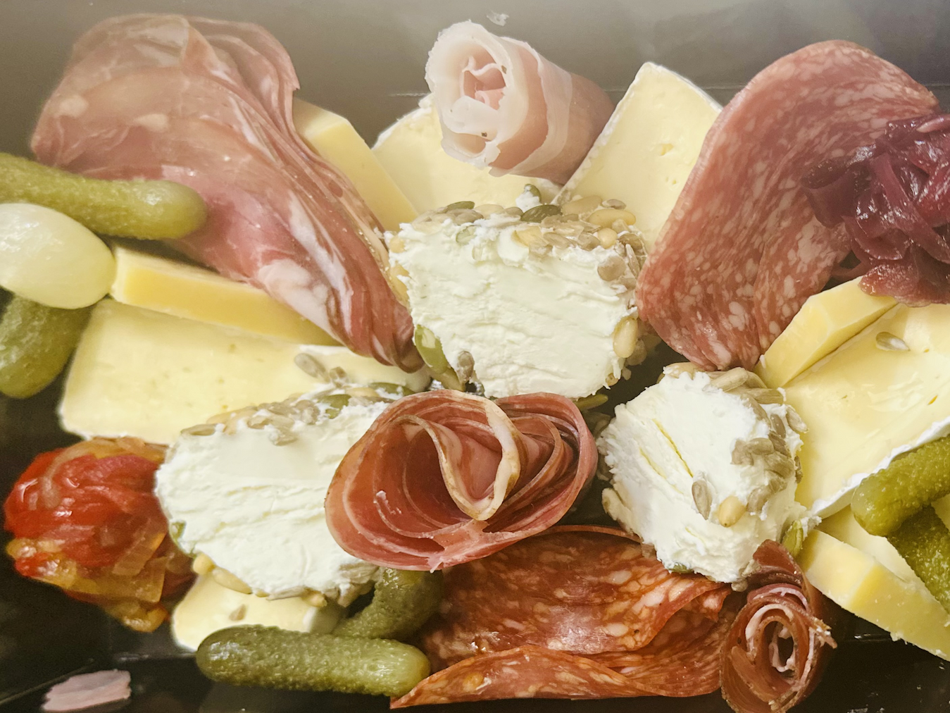 Charcuterie platter with slices of prosciutto and salami, wedges of firm and soft cheeses, seeded goat cheese rounds, cornichon pickles, and a small portion of chutney.