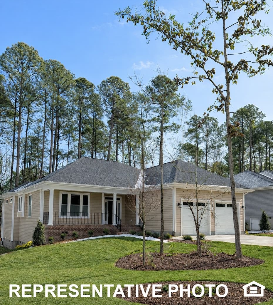 Single-story beige home with front porch and garage, set on a gently sloped green lawn with young trees and tall pines in the background under a clear blue sky.