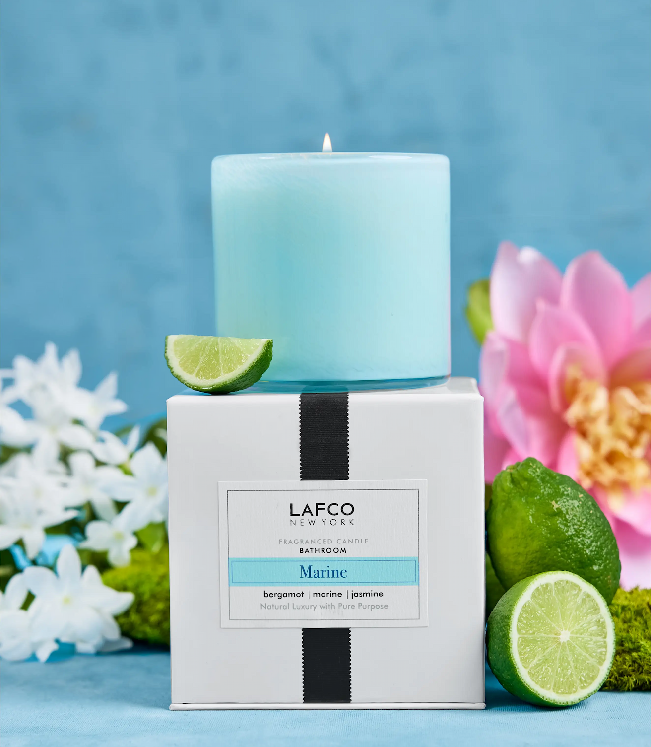 Light blue glass candle on a boxed base labeled “LAFCO New York Marine,” styled with lime halves and flowers against a soft blue background.