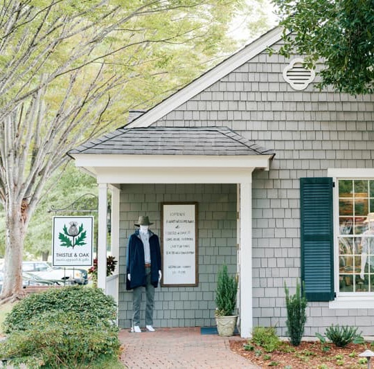 Exterior of a quaint shingled boutique with a small covered porch and green shutters, a Thistle & Oak sign by the walkway, and a mannequin dressed in a jacket and hat standing near the entrance under leafy trees.