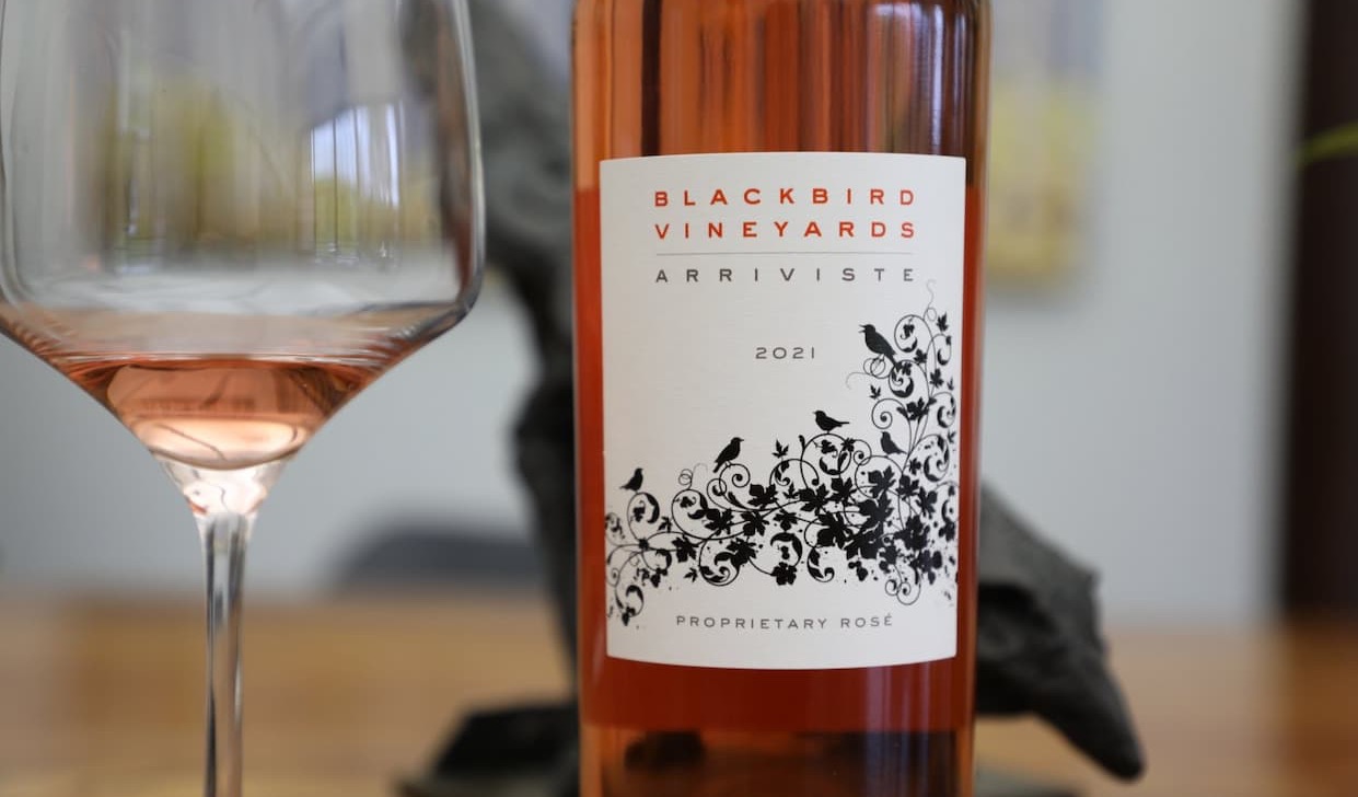 A bottle of Blackbird Vineyards Arriviste Proprietary Rosé 2021 stands beside a partially filled wine glass. The wine’s soft pink hue is reflected in both the bottle and glass, while the label features elegant black floral vines and perched birds against a white background. The close-up composition and blurred background create a refined, sophisticated presentation that highlights the wine’s color and distinctive label design.