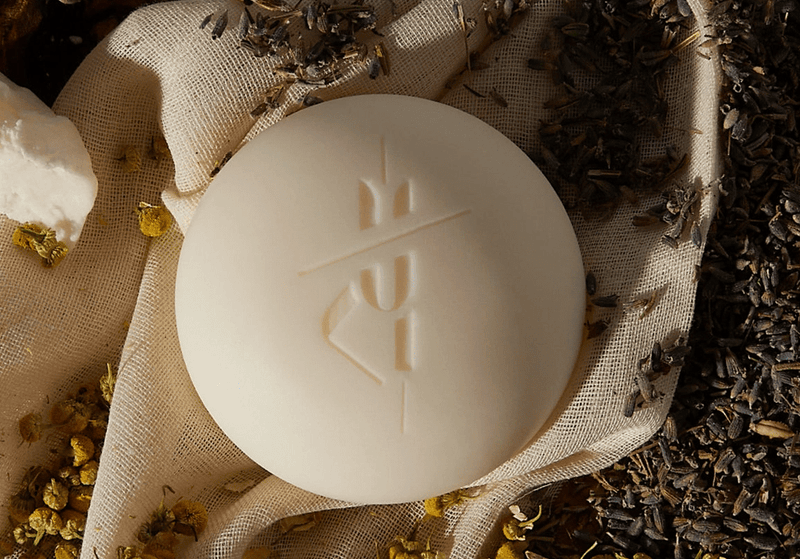 Round cream-colored body bar with an embossed logo resting on natural fabric surrounded by dried lavender and chamomile flowers.