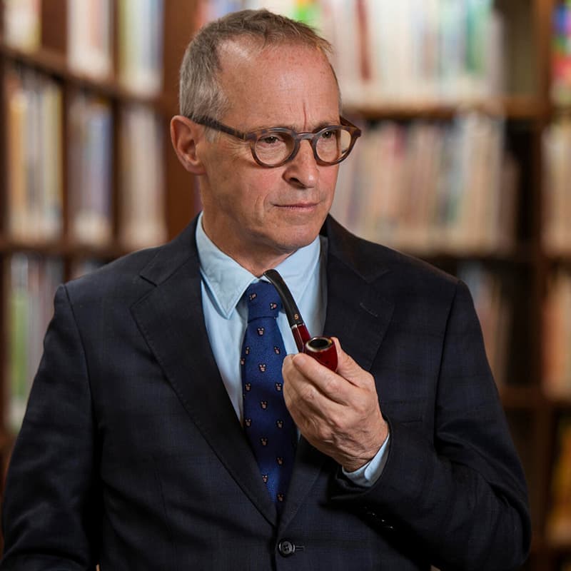 Man in a dark suit and tie stands in front of bookshelves, holding a pipe and looking off to the side with a thoughtful expression. He wears round glasses, giving him a scholarly, contemplative appearance in a library setting.