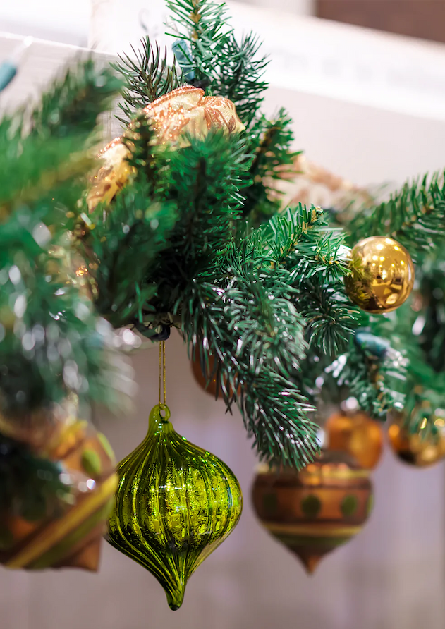 Close-up of a decorated Christmas garland with green pine needles, gold and green ornaments, including a prominent green glass bauble hanging in the foreground. Fearrington Village