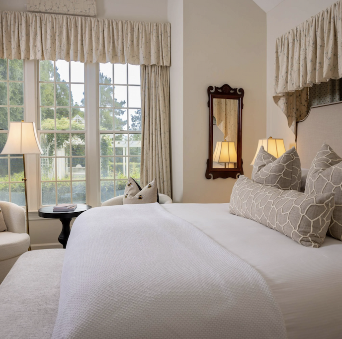 A serene guest room features a large bed dressed in crisp white linens and layered neutral-toned pillows. Tall windows framed with soft patterned drapery fill the space with natural light and offer views of lush greenery outside. A pair of upholstered chairs and a small round table create a cozy sitting area, while warm bedside lamps and classic furnishings add an elegant, inviting feel.