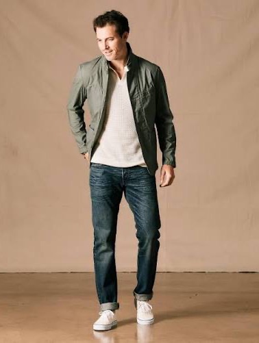 A man models an olive green casual jacket with a white knit shirt, dark jeans and white sneakers. Fearrington Village.