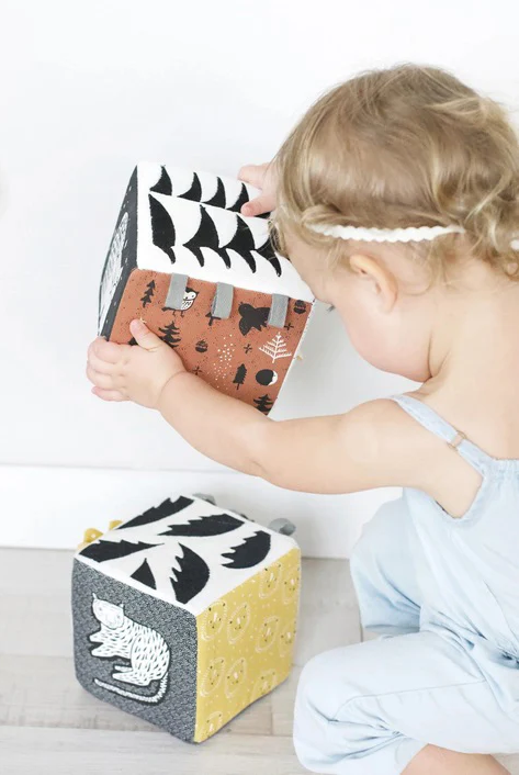 Toddler playing with a soft fabric activity block featuring animal and nature patterns, seated on a light floor.