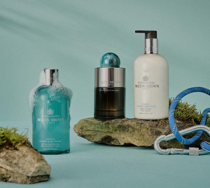 Molton Brown Coastal Cypress & Sea Fennel collection displayed on seaside rocks, featuring eau de toilette, bath & shower gel, and body lotion with ocean spray and coastal greenery.