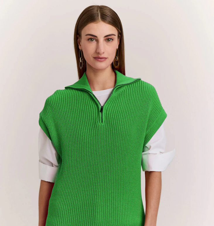 Woman wearing a bright green knit sleeveless zip-neck sweater layered over a white shirt, standing against a neutral background.