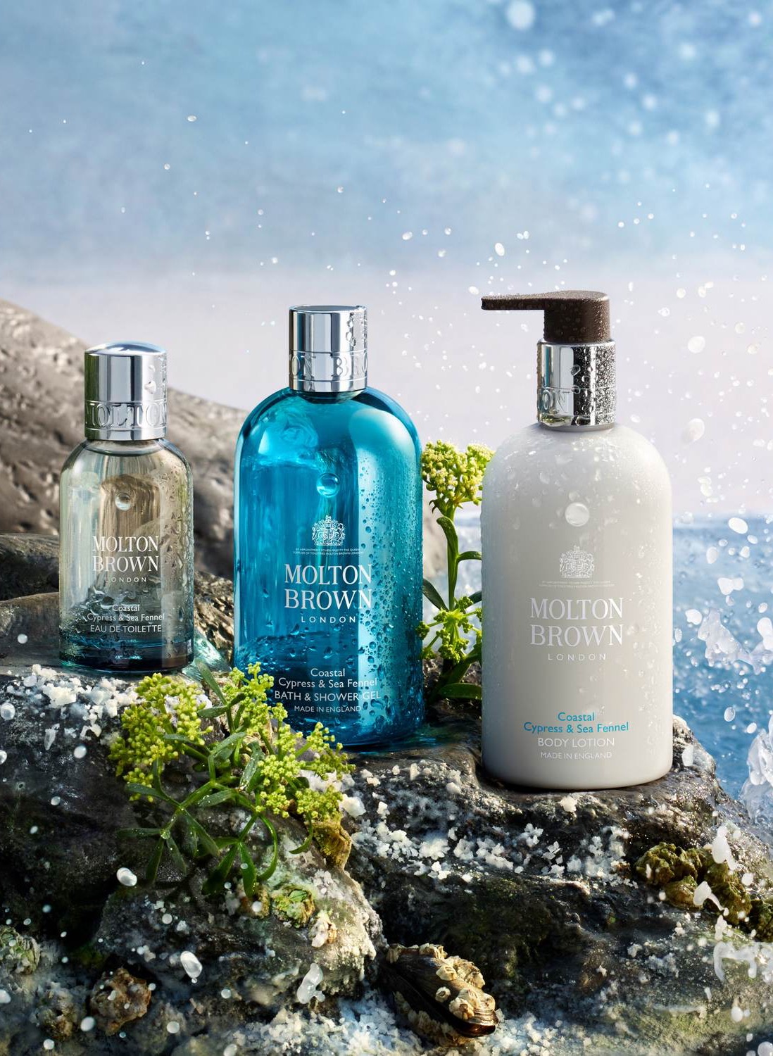 Molton Brown bath and body products arranged on seaside rocks, including a blue bath and shower gel, a white body lotion with a pump, and a smaller fragrance bottle, surrounded by sea spray, greenery, and ocean waves in the background.