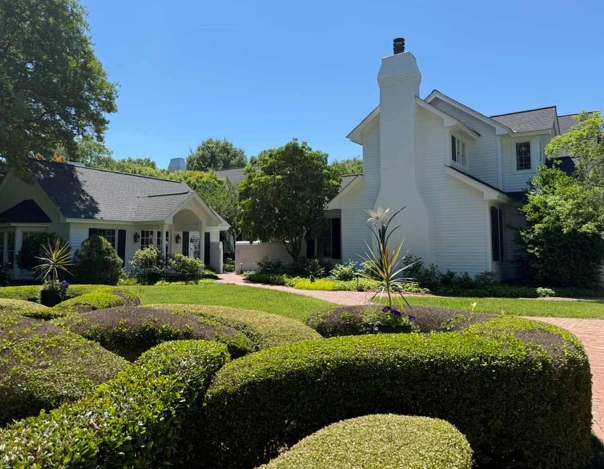 White cottage-style homes with dark shutters sit around a manicured garden of sculpted hedges and curved pathways, under a clear blue sky with lush trees framing the scene.
