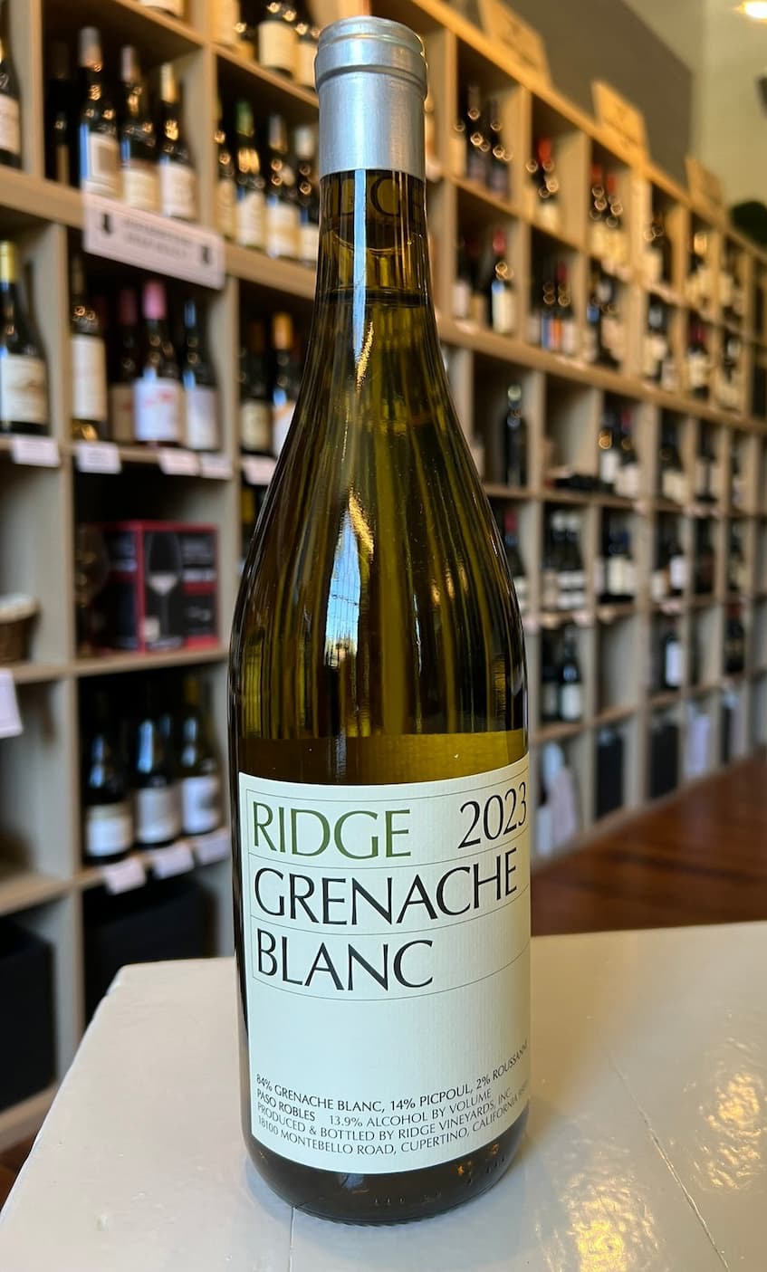 A bottle of 2023 Ridge Grenache Blanc displayed on a counter inside a wine shop lined with shelves of bottles.
