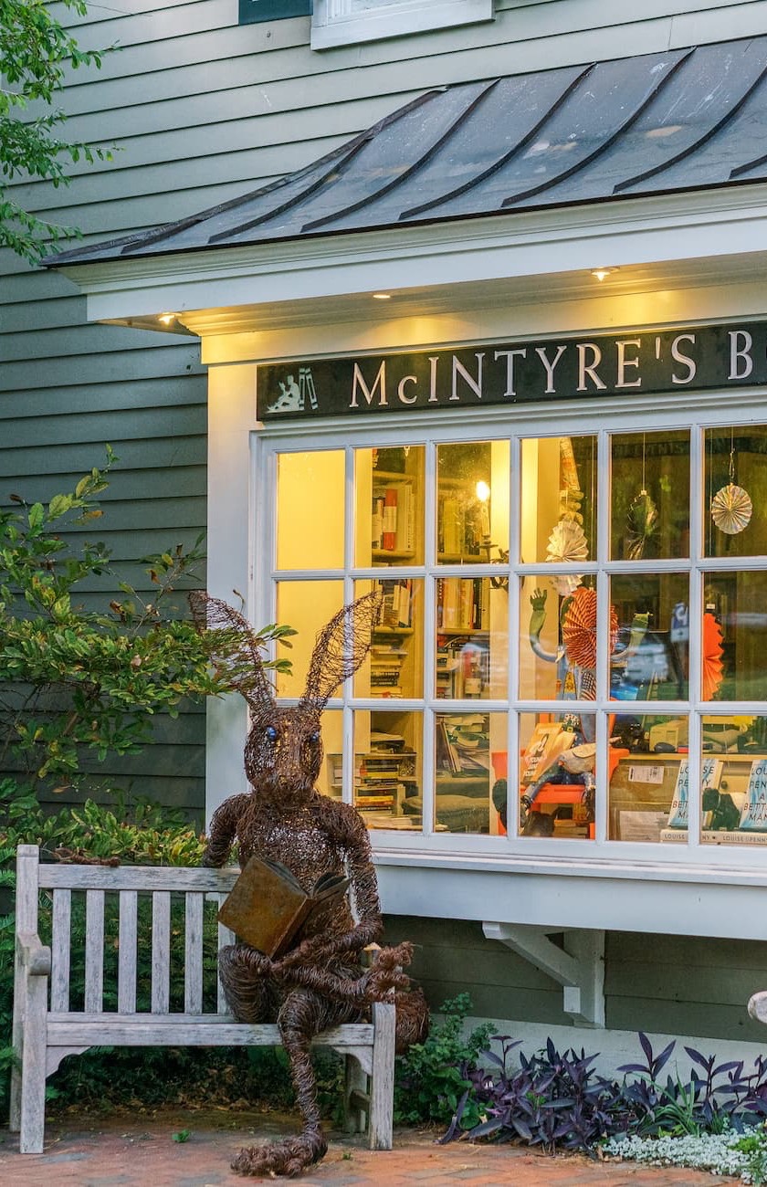 Exterior of McIntyre's Books at dusk, with warm lights glowing through the windows and a whimsical wire rabbit sculpture reading a book on a bench outside.